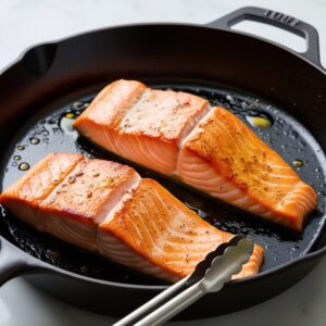 Two salmon fillets cooking in a cast-iron skillet. The skin is turning golden-brown and crispy. Olive oil sizzles in the pan. Tongs are visible nearby. The salmon flesh is becoming lighter pink as it cooks.